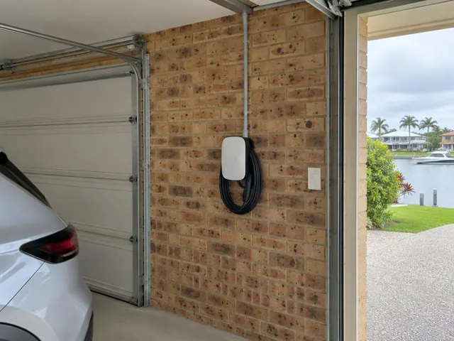 Wall-mounted EV charger with coiled cable on a brick garage wall beside a roller door in Broadbeach Waters